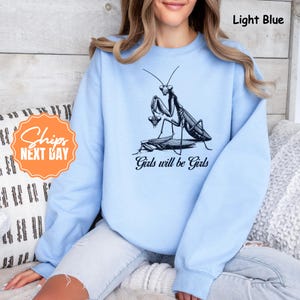 May include: Light blue sweatshirt with a black and white graphic of a praying mantis and the text "Girls will be Girls". The sweatshirt is a crew neck style.