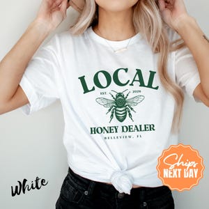 May include: White t-shirt with a green graphic that reads "LOCAL HONEY DEALER" with a bee illustration. The text "BELLEVIEW, FL" is below the bee. The shirt also has the text "EST. 2026".