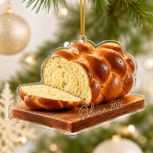 May include: A clear acrylic Christmas ornament shaped like a loaf of braided bread on a wooden cutting board. The bread is golden brown with a visible crumb texture. The text Olivia 2026 is printed in white script on the board. It hangs by a gold cord.