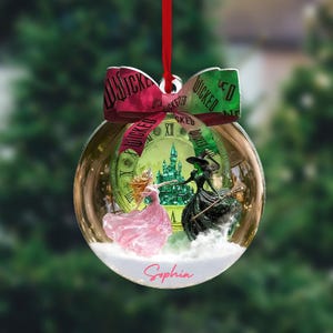 May include: A clear glass Christmas bauble with a red ribbon. Inside, a scene from the musical "Wicked" features a princess in a pink dress and a green witch. The bauble has a green clock face background and the name "Sophia".