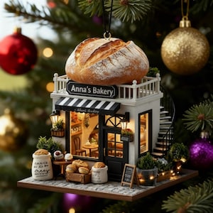 May include: A Christmas tree ornament shaped like a bakery, with a loaf of bread on top. The bakery has a black and white striped awning and the text "Anna's Bakery". The ornament includes miniature baked goods and a spiral staircase.