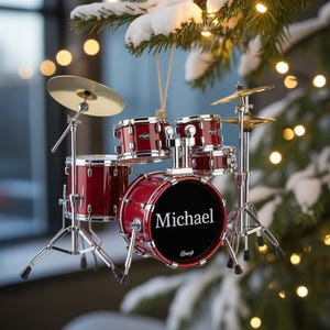 May include: Miniature red drum kit Christmas ornament with the name "Michael" on the bass drum. The ornament includes cymbals and stands, hanging from a gold string. The ornament is displayed on a snow-covered Christmas tree.