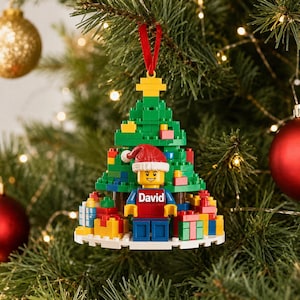 May include: A Christmas tree ornament constructed from plastic building blocks featuring a green tree, a yellow star topper, and a minifigure wearing a red Santa hat. The minifigure wears a red shirt with the name David printed in white text.