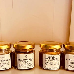 May include: Six small glass jars with gold lids, each containing a different type of honey. The jars are labeled with the honey's name and ingredients. The labels are white with black text. The jars are arranged in a row.