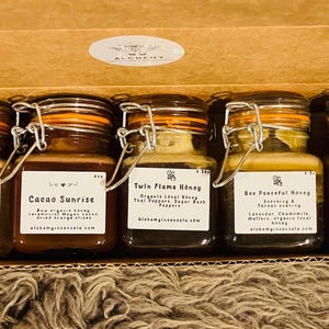 May include: A gift set of five small glass jars filled with honey, each labeled with a different flavor. The jars are in a cardboard box. The labels include text such as "Sweet Cinnilla" and "Cacao Sunrise".