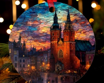 May include: Round Christmas ornament featuring a stained-glass style depiction of a European city square at dusk. The ornament has a red ribbon for hanging. The artwork showcases a cathedral with tall spires and a vibrant sunset.