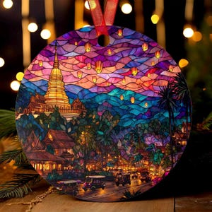 May include: Round Christmas ornament featuring a colorful stained-glass style illustration of a temple, buildings, and tuk-tuks. The ornament has a red ribbon for hanging. The artwork depicts a vibrant sunset with floating lanterns.