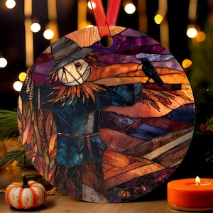 May include: Round ornament with a stained-glass design featuring a scarecrow and a crow against a sunset backdrop. The scarecrow has a straw-filled head and a blue coat. A red ribbon is attached for hanging. The ornament is a fall or Halloween decoration.