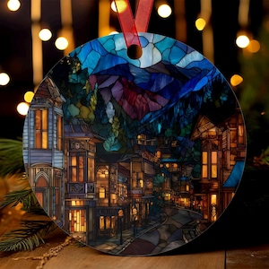 Peut inclure: Ornement de Noël rond avec un motif de vitrail représentant une ville dans une vallée montagneuse. L'œuvre d'art montre des bâtiments aux fenêtres éclairées, une rue sinueuse et un ciel nocturne aux teintes bleues et violettes. Un ruban rouge est attaché pour l'accrocher.