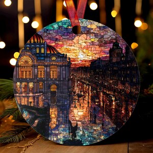 May include: Circular Christmas decoration with a stained-glass effect depicting a city street at sunset. The ornament has a red ribbon for hanging. The artwork shows buildings, streetlights, and figures, with a vibrant, colourful sky.