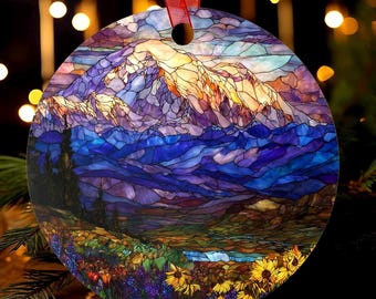 May include: Round Christmas ornament with a stained-glass design depicting a mountain landscape. The ornament features a red ribbon for hanging. The artwork includes shades of blue, purple, yellow, and green, with a detailed mountain range and floral elements.