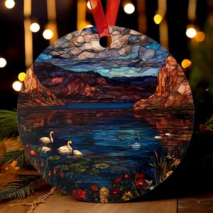 Könnte beinhalten: Runder Ornament mit einer Landschaft im Glasmalerei-Stil, mit See, Bergen und Schwänen. Das Ornament hat ein rotes Band zum Aufhängen. Das Kunstwerk verwendet Blau-, Braun- und Orangetöne.