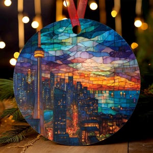 May include: Round Christmas ornament featuring a stained-glass style depiction of the Toronto skyline at sunset. The CN Tower is prominent, with a red ribbon for hanging. The ornament has a colorful, mosaic-like design with shades of blue, orange, and yellow.