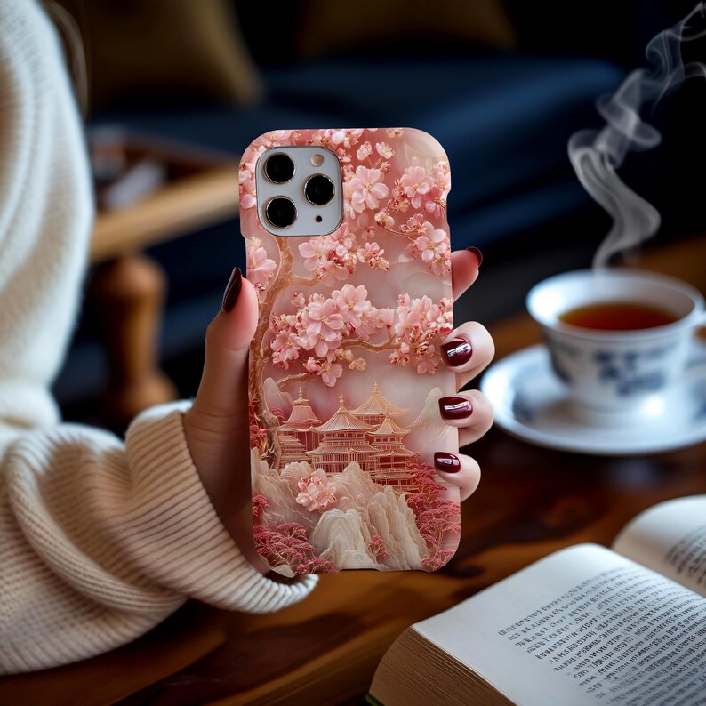 May include: A pink phone case featuring a detailed illustration of a Japanese pagoda scene with cherry blossoms. The case has a 3D effect, with the blossoms and buildings slightly raised. The color palette includes shades of pink, white, and brown.