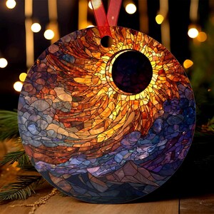Op de afbeelding: Ronde kerstversiering met een glas-in-lood ontwerp van een zonsverduistering. De versiering heeft een rood lint om op te hangen. Het ontwerp bevat tinten oranje, geel, blauw en paars, met een donkere cirkel die de maan voorstelt.