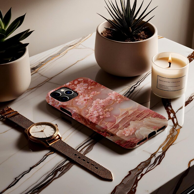 May include: A floral iPhone case with a pink and white design, a gold watch with a mesh band, and a lit candle sit on a marble surface. Two small potted plants are also present. The phone case features a cherry blossom pattern.