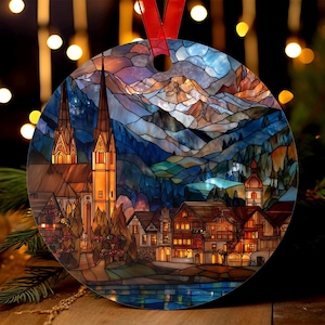 May include: Circular Christmas decoration featuring a stained glass effect of a European village with mountains, a lake, and church spires. It has a red ribbon for hanging. The colour scheme includes blues, oranges, and browns.