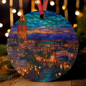 Op de afbeelding: Ronde kerstversiering met een kleurrijk glas-in-loodontwerp van een stadsgezicht met een hoge toren. De versiering heeft een rood lint om op te hangen. Het kunstwerk toont gebouwen, bomen en lantaarns in blauwe, oranje en paarse tinten.