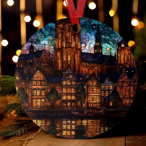 May include: Round Christmas ornament featuring a colorful illustration of a European cityscape at night. The design includes buildings, a church, and a river with reflections. The ornament has a red ribbon for hanging.