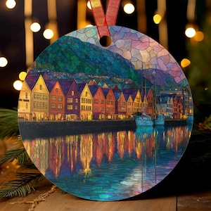 Puede incluir: Adorno navideño redondo con una colorida representación de un paisaje costero al estilo vidriera, con edificios, barcos y montañas de fondo. El adorno tiene una cinta roja para colgar.