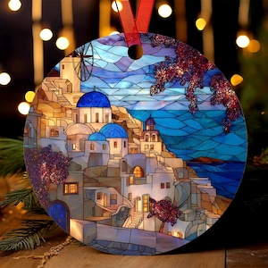 Op de afbeelding: Ronde kerstversiering met een afbeelding in glas-in-loodstijl van een kustdorp met witte gebouwen, blauwe koepels en een windmolen. Het ornament heeft een rood lint om op te hangen en een vage achtergrond.