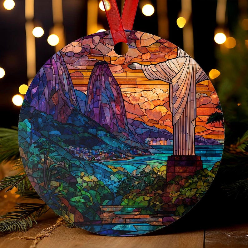 May include: A round Christmas ornament featuring a stained-glass style depiction of Christ the Redeemer statue in Rio de Janeiro. The ornament has a red ribbon for hanging and showcases vibrant colors of blue, purple, orange, and green.