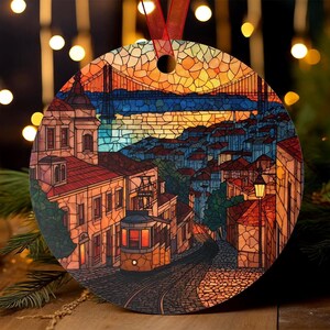 Puede incluir: Adorno navideño redondo con una colorida ilustración estilo vidriera de una ciudad europea con un puente, edificios y un tranvía. El adorno tiene una cinta roja para colgar.
