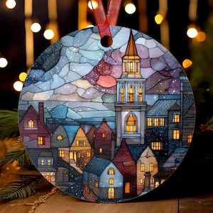 Puede incluir: Adorno navideño redondo con una representación de un pueblo al estilo vidriera. El diseño incluye una iglesia con una torre alta, casas con ventanas iluminadas y un cielo colorido. Se adjunta una cinta roja para colgar.