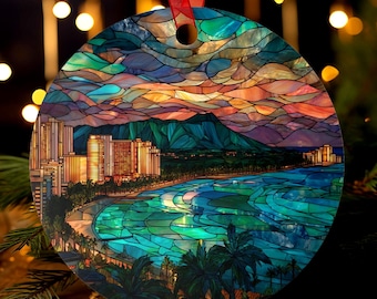May include: Round Christmas ornament featuring a stained-glass design of a coastal city at sunset. The ornament has a red ribbon for hanging. The image shows a beach, palm trees, buildings, and mountains, with a colorful sky.