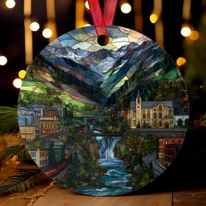 May include: Round Christmas ornament featuring a stained-glass style depiction of a mountain town with a river, waterfall, and train. The ornament has a red ribbon for hanging. The artwork includes buildings, trees, and mountains in shades of green, blue, and brown.