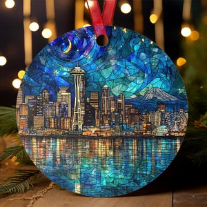 May include: Round Christmas ornament featuring a stained-glass style depiction of the Seattle skyline at night. The ornament includes the Space Needle, a Ferris wheel, and a mountain range, with a red ribbon for hanging.