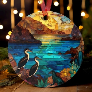 May include: Circular ornament with a stained-glass effect depicting a coastal landscape. Two black and white birds are perched on rocks near blue water. The background shows a sunset over the water and mountains. A red ribbon is attached for hanging.