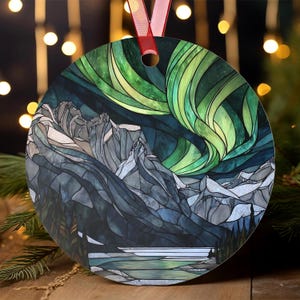 May include: Round Christmas ornament featuring a stained-glass style depiction of the Northern Lights over a mountain range. The ornament has a red ribbon for hanging. The design incorporates shades of green, blue, gray, and white.