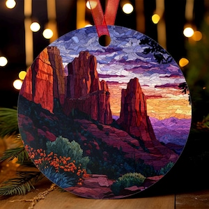 May include: Round Christmas ornament featuring a Southwestern landscape with red rock formations and a colorful sunset. The ornament has a red ribbon for hanging and is decorated with a detailed, stained-glass style design.
