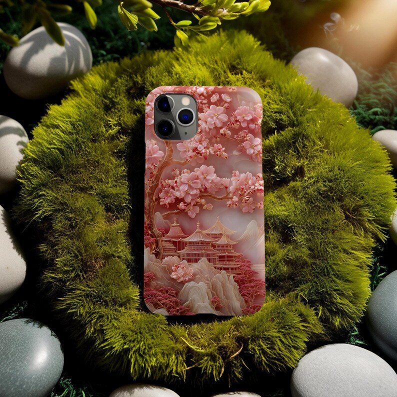May include: A phone case featuring a pink cherry blossom design with a Japanese pagoda scene. The case has a textured appearance and a raised relief of the blossoms and buildings. The case is set against a background of green moss and smooth stones.