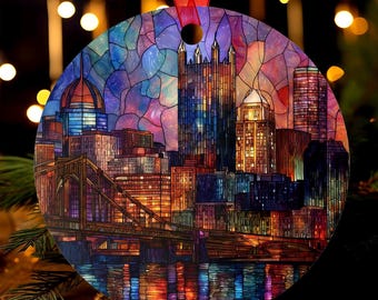 May include: Round Christmas ornament featuring a colorful stained-glass depiction of a city skyline with a bridge and water reflection. The ornament has a red ribbon for hanging. The buildings are illuminated with warm colors.