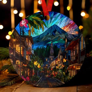May include: Round Christmas ornament featuring a stained-glass design of a town street scene with a mountain backdrop. The ornament has a red ribbon for hanging. The artwork includes buildings, trees, and a road, with warm lighting.