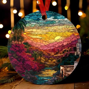 May include: Round Christmas ornament featuring a stained-glass design of a sunset over a landscape with mountains, palm trees, and a river. The ornament has a red ribbon for hanging. The color palette includes shades of blue, orange, pink, and green.
