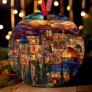 May include: Round Christmas ornament featuring a stained-glass style depiction of a city with domes and buildings. The ornament has a red ribbon for hanging. The color palette includes shades of orange, blue, purple, and gold, with a sunset-like sky.
