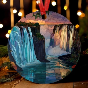 May include: A round ornament featuring a colorful illustration of a waterfall scene. The artwork depicts cascading water, cliffs, and a sunset sky. A red ribbon is attached for hanging. The ornament is likely for holiday or decorative use.