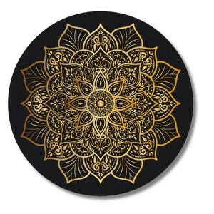May include: A round, black decorative item featuring a gold mandala design. The mandala has an intricate floral pattern with detailed swirls and leaf-like shapes. The design is centered and symmetrical, with a golden center.