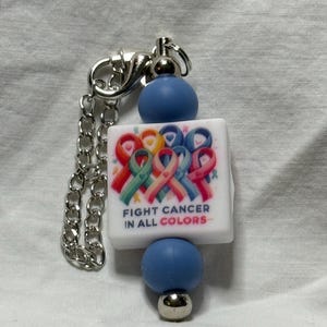 Puede incluir: Un llavero de cuentas con una cadena y un cierre plateados. El diseño incluye un cuadrado blanco con cintas de concienciación sobre el cáncer de colores y el texto "FIGHT CANCER IN ALL COLORS". Hay cuentas azules en la parte superior e inferior.