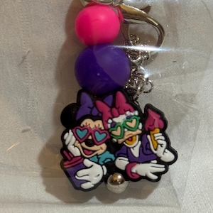 Puede incluir: Un llavero con Minnie Mouse y Daisy Duck con gafas de sol y helados. El dije está unido a una cadena con cuentas rosas y moradas y un cierre plateado.