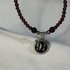 May include: A burgundy beaded necklace with a silver charm. The charm is a circular design with the words "PLATFORM 9 3/4" and the numbers 9, 3, and 4. The necklace has black and iridescent beads.