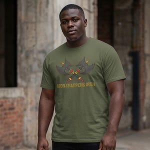 May include: Olive green t-shirt featuring a graphic of two crossed axes with red, yellow, and green accents. The text "IRON SHARPENS IRON" is printed below the axes in a gold color. The shirt is being worn.