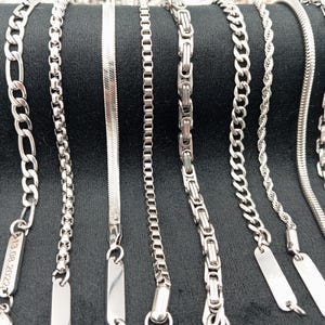 May include: A collection of silver chains with different link styles and textures. The chains are displayed on a black background and have small rectangular tags attached to them.