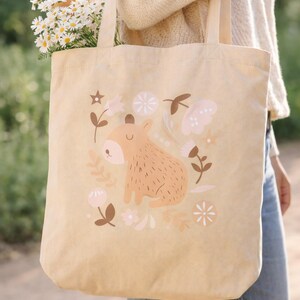 May include: Beige canvas tote bag featuring a whimsical illustration of a capybara surrounded by flowers and leaves in soft pink and brown tones. The bag is filled with daisies and a baguette.