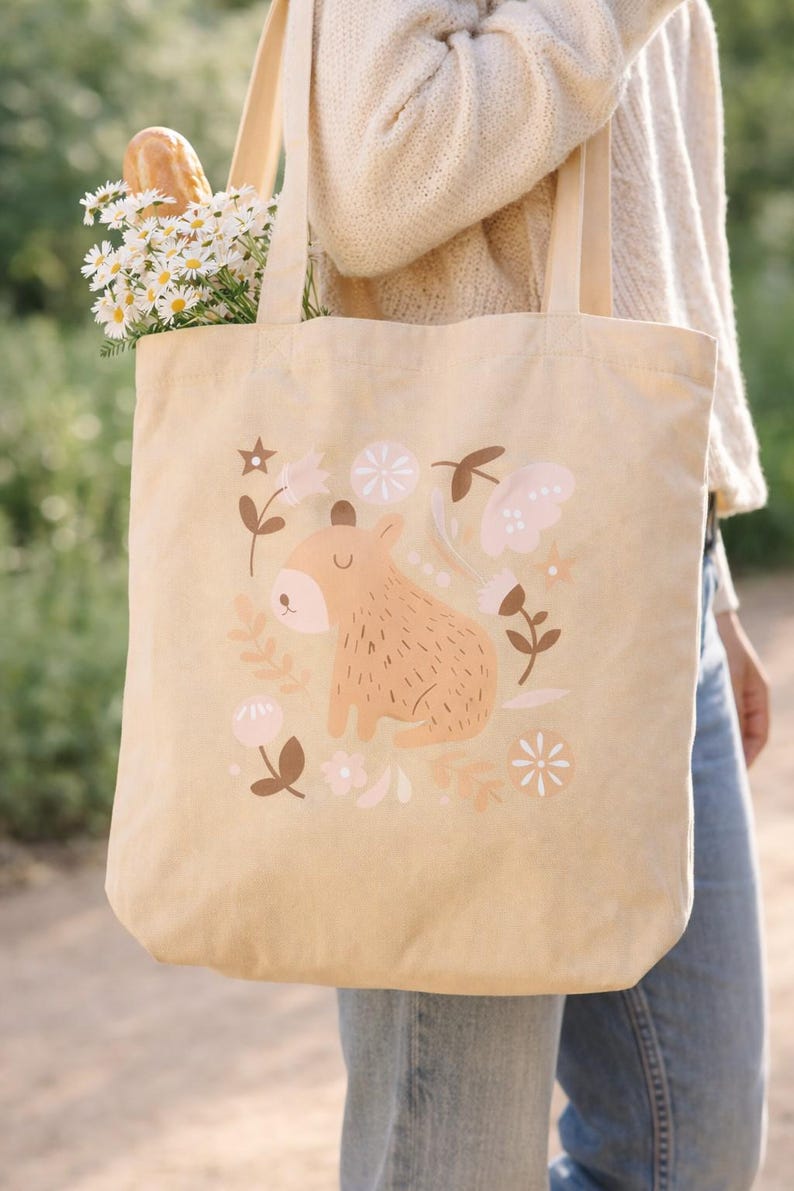 May include: Beige canvas tote bag featuring a whimsical illustration of a capybara surrounded by flowers and leaves in soft pink and brown tones. The bag is filled with daisies and a baguette.