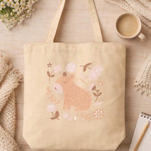 May include: Beige canvas tote bag featuring a capybara illustration surrounded by flowers in shades of pink and brown. The bag has a simple design with long handles, perfect for carrying items. A cozy, neutral-toned aesthetic.