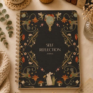 Puede incluir: Un diario negro con encuadernación en espiral dorada, con el texto "SELF REFLECTION JOURNAL". La portada está adornada con ilustraciones florales y animales, incluyendo colibríes y un conejo. El diario está rodeado de una vela, libros y una taza de té.